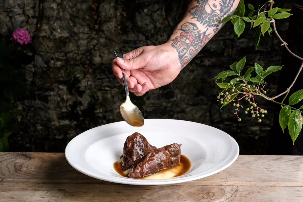 Mano sujetando una cuchara mientras vierte una salsa sobre un planto de carne